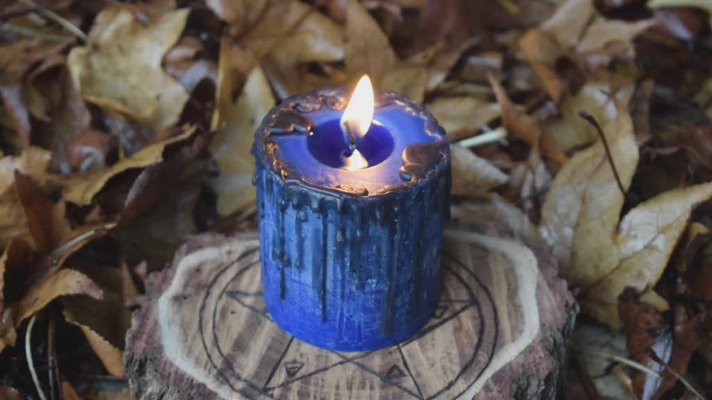 A blue pillar candle with black drips sits with a flame licking from its wick on a timber pentagram disk nestled on a bed of leaves.