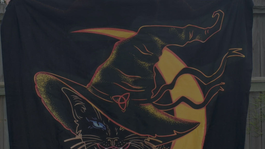 Person unrolling black hand painted fabric throw depicting a black cat wearing witches hat outlined in yellow and orange with yellow crescent moon in background