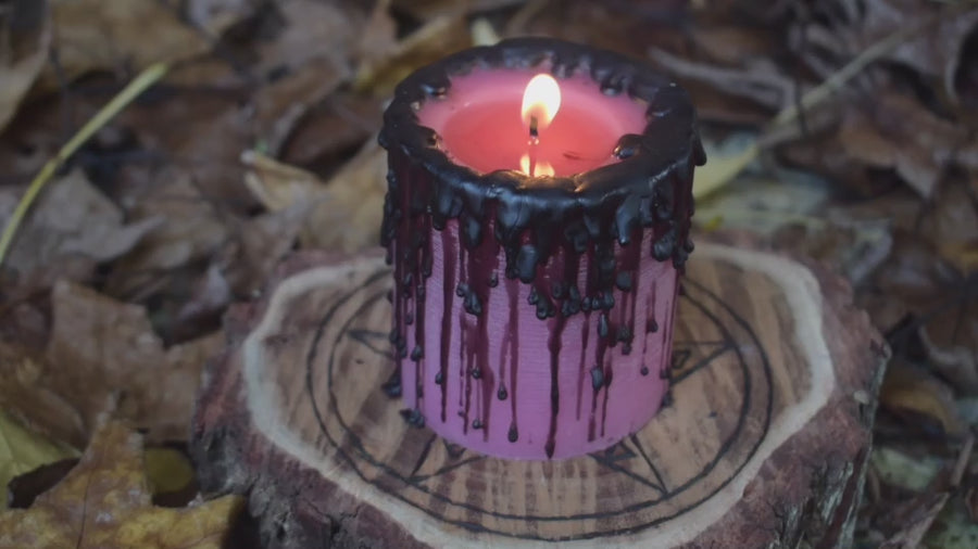 A lit pink pillar candle with a flame licking from its wick sits on a pentagram disk nestled on a bed of autumn leaves