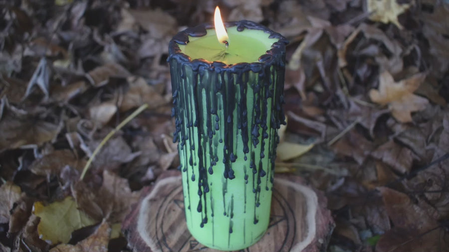 A green pillar candle with black drips with its wick flickering alight nestled on a bed of autumn leaves.