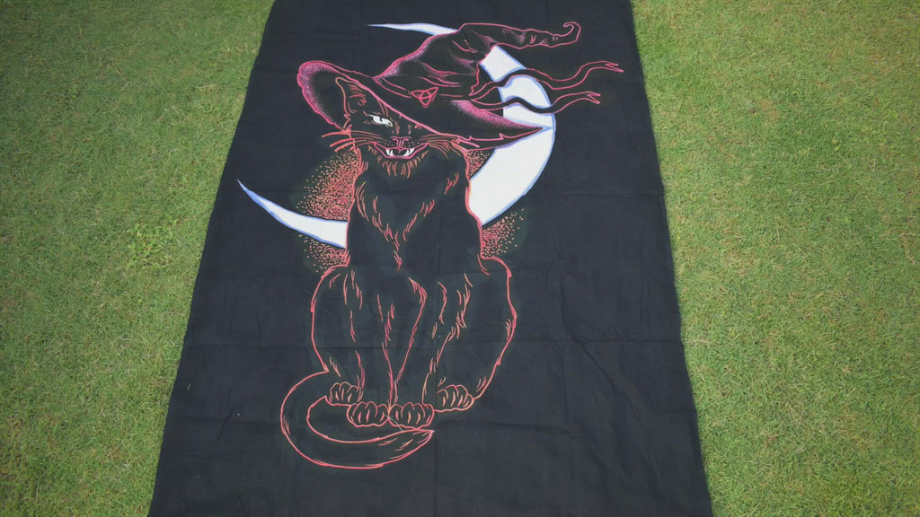 Person unrolling a hand painted black fabric throw depicting a black cat wearing witches hat outlined in pink and orange with white crescent moon in background