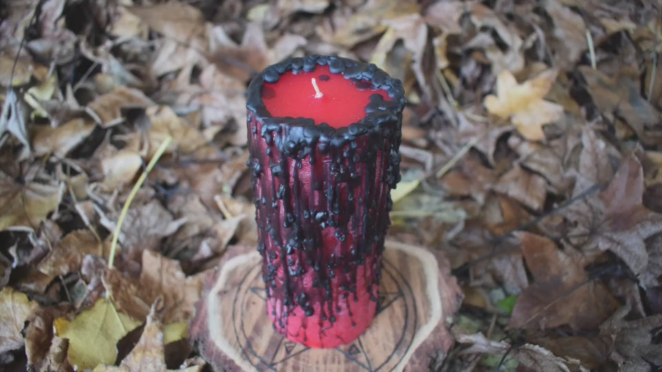 A red pillar candle with black wax drips with a flame dancing from its wick rests on a pentagram disk nestled on a bed of autumn leaves