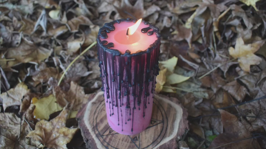 A flame flickers from a pink pillar candle with black drips as it rests on a bed of autumn leaves
