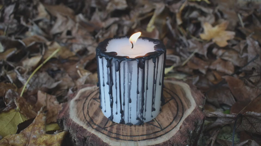 A black and white pillar candle with a flame flickering from its wick rests on a wooden pentagram disk on a bed of autumn leaves
