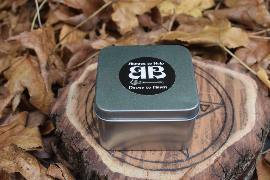 A tin with an always to help never to harm BB sticker on the top of the lid resting on a piece of timber nestled in a bed of leaves