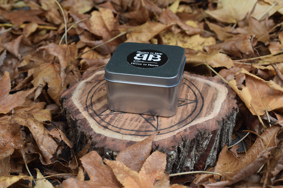 A tin with an always to help never to harm BB sticker on the top of the lid resting on a piece of timber with a pentagram on it nestled in a bed of leaves