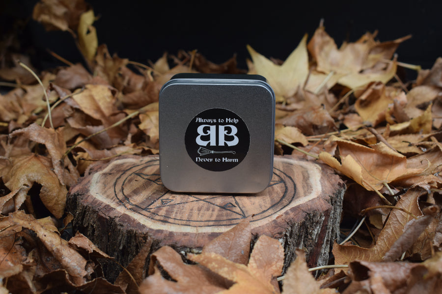 A tin with an always to help never to harm BB sticker on the top of the lid sitting on a piece of timber with a pentagram burnt on it nestled on a bed of leaves