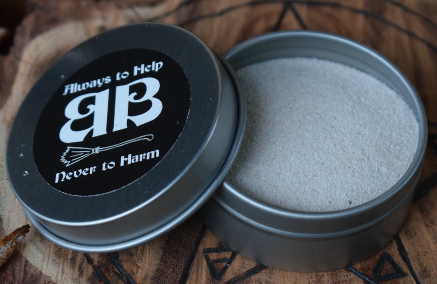 An open tin with an always to help never to harm BB sticker on the top of the lid showing the contents of white sand sitting on a piece of wood with a pentagram on it 