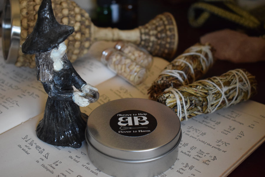 A tin of sand with label on lid saying always to help never to harm BB on the lid resting on a page of hieroglyphics with a ceramic statue of a witch standing over it with smudge sticks, vials of frankincense and a goblet in the background