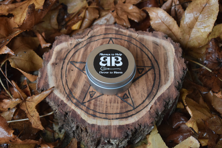 A tin with an always to help never to harm BB sticker on the top of the lid on a piece of wood with a pentagram on it nestled in a bed of leaves