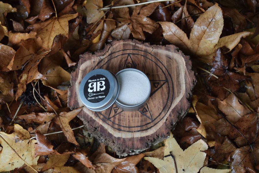 An open tin with an always to help never to harm BB sticker on the top of the lid showing the contents of white sand sitting on a piece of wood with a pentagram on it nestled in a bed of leaves