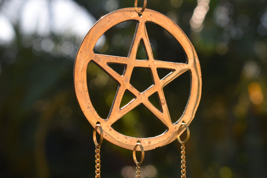 A bronze pentagram pentacle metal wind chime hanging in the garden with mottled greenery in the background
