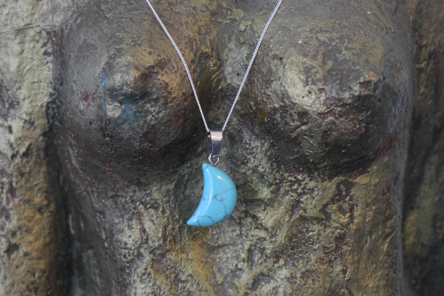 A blue howlite crystal crescent moon necklace on a sterling silver chain hanging on the neck of a bronze goddess sculpture