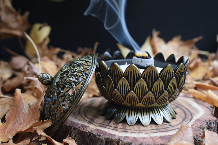 A metal lotus trinket box with a lid to the side while smoke wafts from its heart resting on a wooden disk with autumn leaves in the background