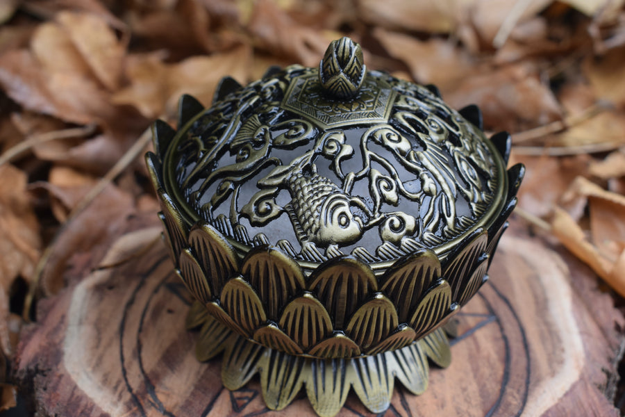A metal lotus trinket box with a lid resting on a wooden pentagram disk with a bed of leaves in the backgroud
