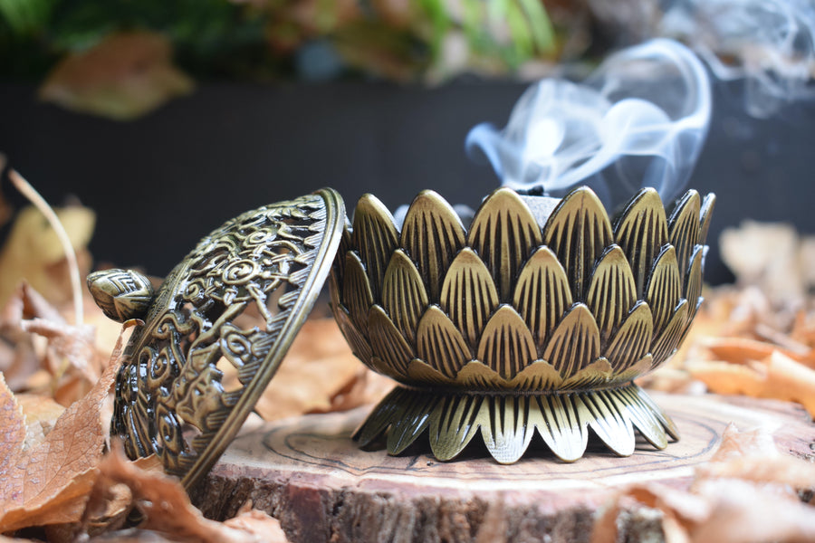 A metal lotus trinket box with a lid to the side while smoke wafts from its heart.  
