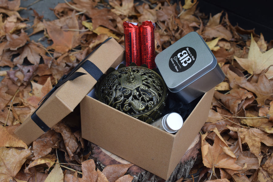 A ribboned box with an open lid showing an metal cauldron, two sticks of self-lighting charcoal disks, a tin of sand and a glass vial of frankincense resin resting on a bed of autumn leaves