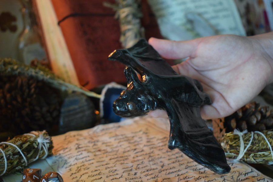 A handmade ceramic flying fox bat being held by a hand with herbs and crystals in the background