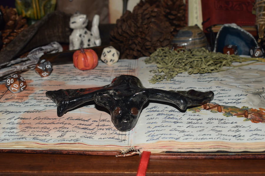 A handmade ceramic flying fox bat resting on an open grimoire with herbs, dice and crystals in the background