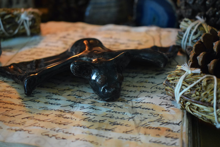 A handmade ceramic flying fox bat resting on an open grimoire with herbs and crystals in the background