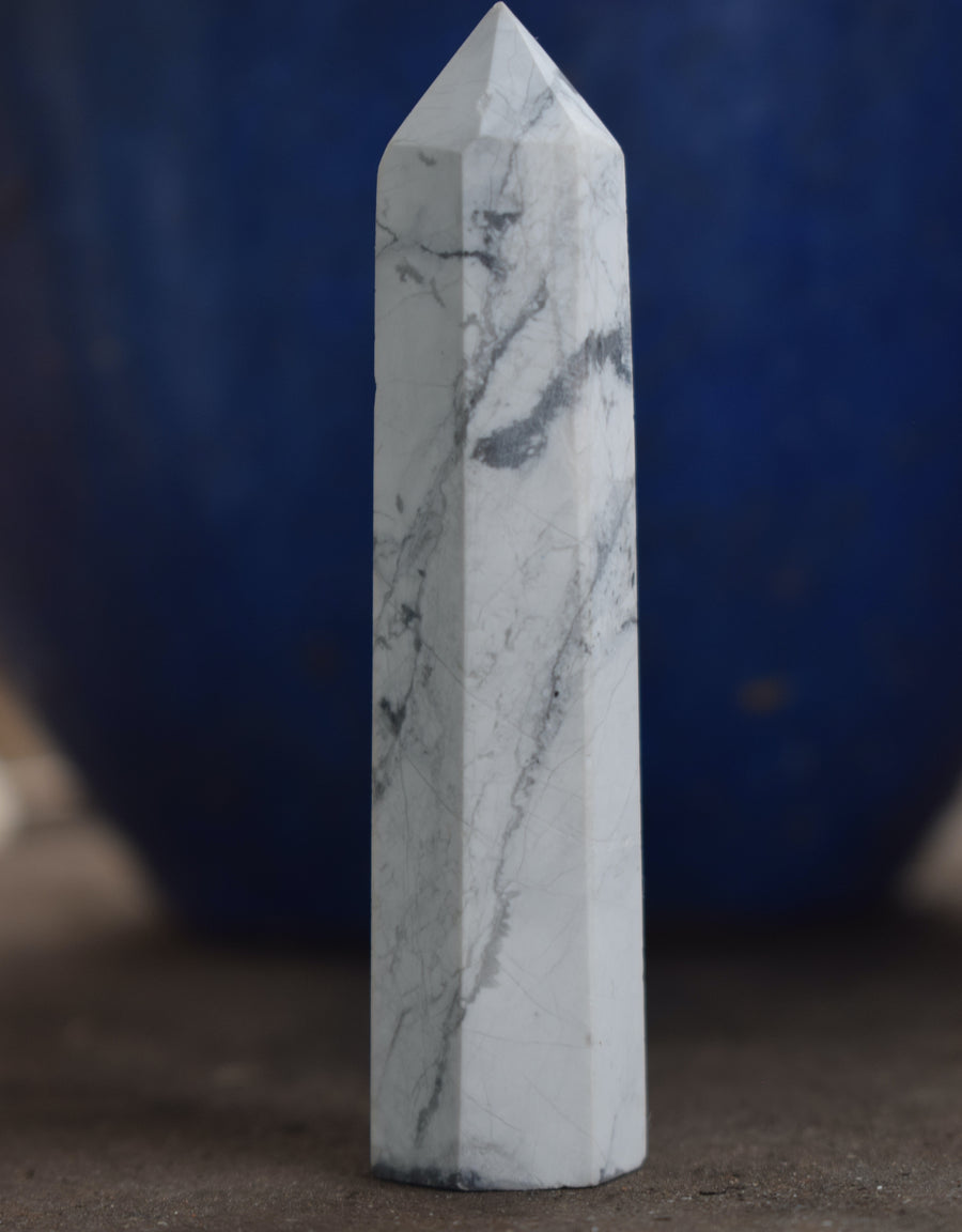 A white howlite obelisk crystal point with a blue background.