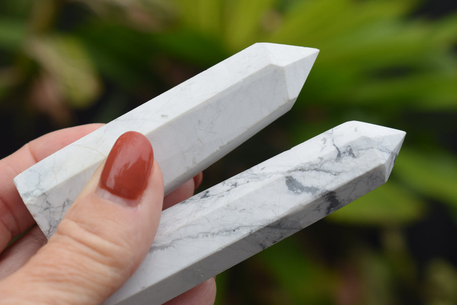 A hand holding two white howlite obelisk crystal points resting with greenery in the background.