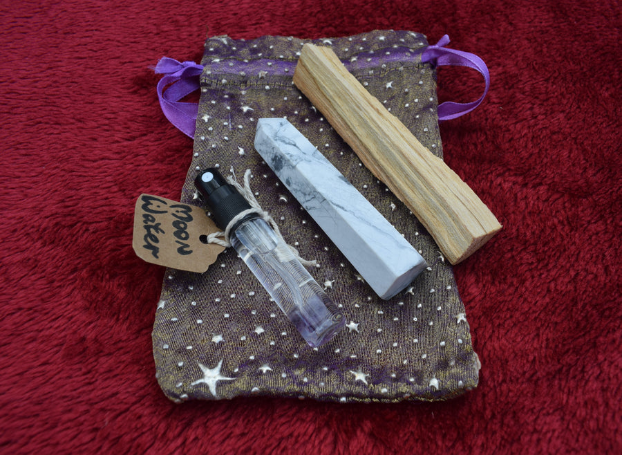A white howlite crystal point with a palo santo stick, a glass spray bottle of moon water resting on a purple and gold drawstring bag.