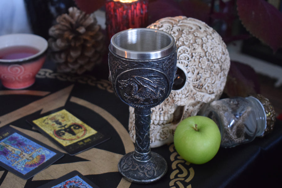 A goblet with a 3d black dragon on it rests on an altar with snake skin, a skull and tarot cards