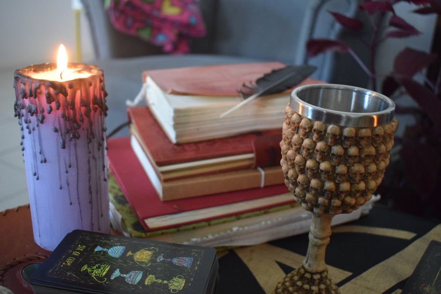 A goblet with skulls all over it sits on an altar with books, a candle, tarot cards and a black feather