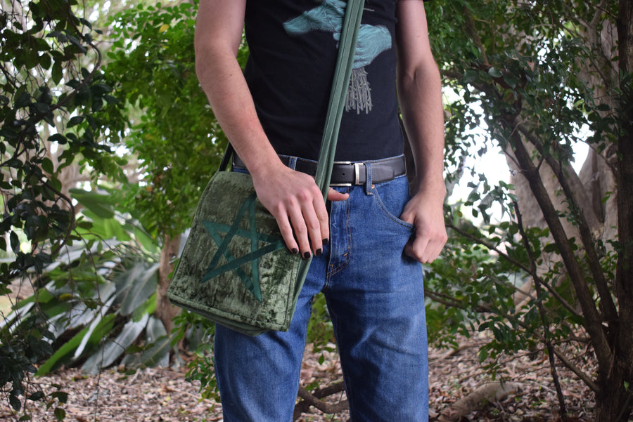 Person standing in the forest wearing a green velvet shoulder bag with pentagram on the front