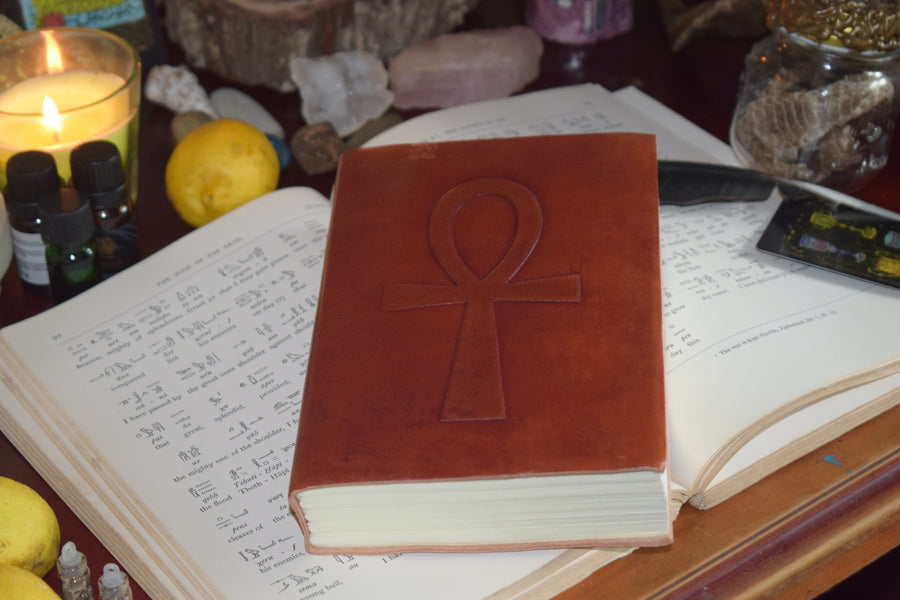 A leather-bound book with an ankh embossed on the front sits on an open page of hieroglyphs from the Egyptian Book of the Dead surrounded by crystals, candles, snakeskin and a feather