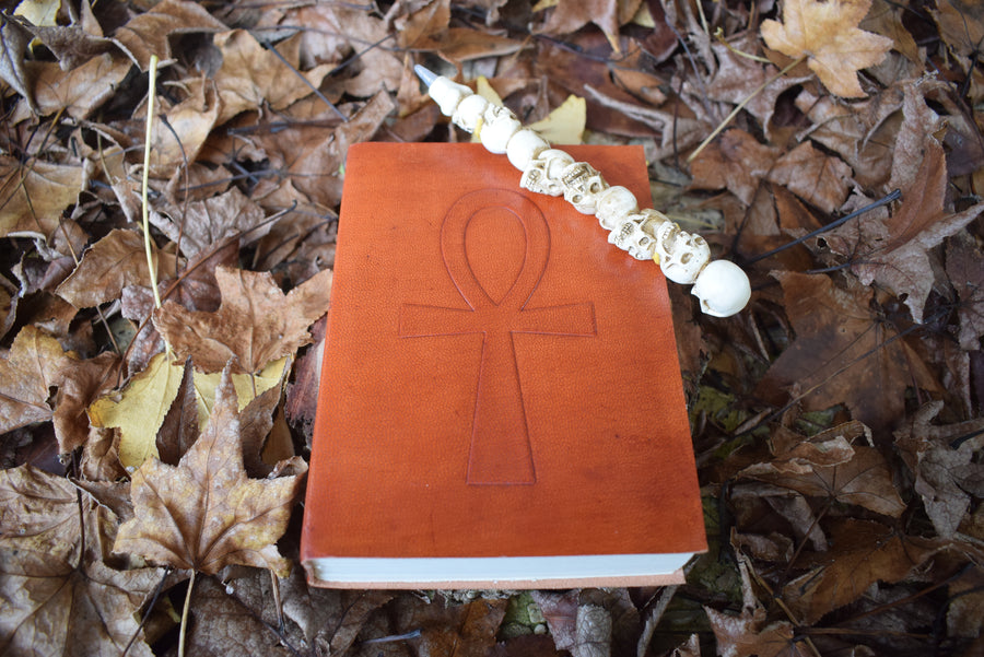 A leather-bound book with an ankh embossed on the front sits on bed of autumn leaves with a pen of skulls resting on it