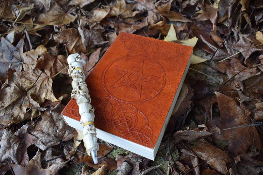A leather-bound book of shadows with a pentagram pentacle and celtic knots embossed on the cover rests in a bed of autumn leaves with a pen of skulls on top.