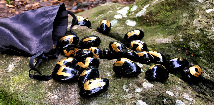 Black runes with gold symbols spilled out from a black drawstring pouch onto a mossy rock