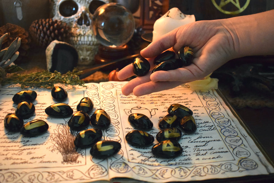 Black runes with gold symbols spilled out by a hand onto an open page of a grimoire with herbs, flowers and crystals in the background