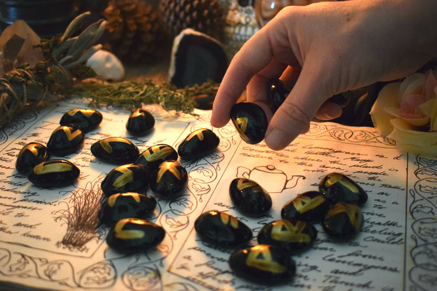 Black runes with gold symbols spilled out onto an open page of a grimoire with a hand picking up two runes with herbs, flowers and crystals in the background