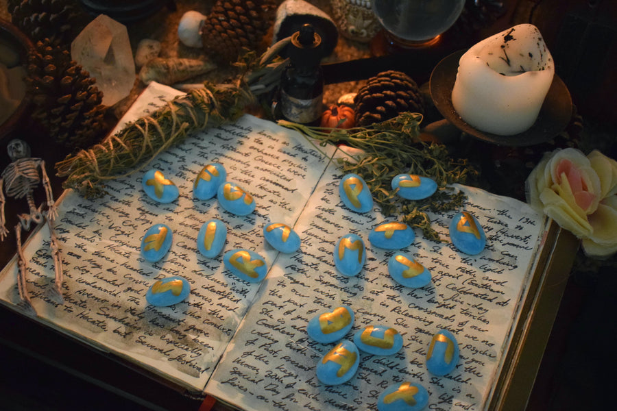 A set of handmade blue elder futhark runes resting on a grimoire surrounded by herbs, candles, crystals on an altar