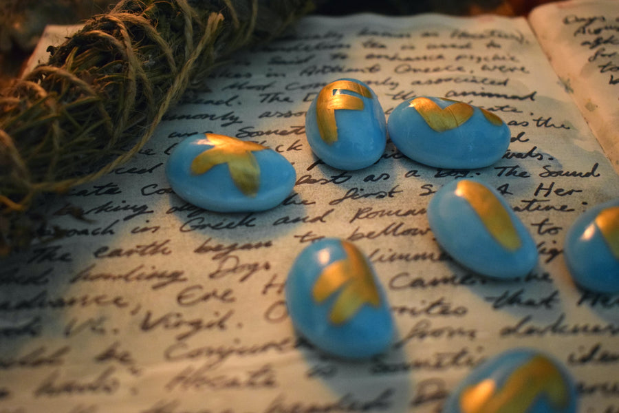 A set of handmade blue elder futhark runes resting on a grimoire surrounded by herbs on an altar