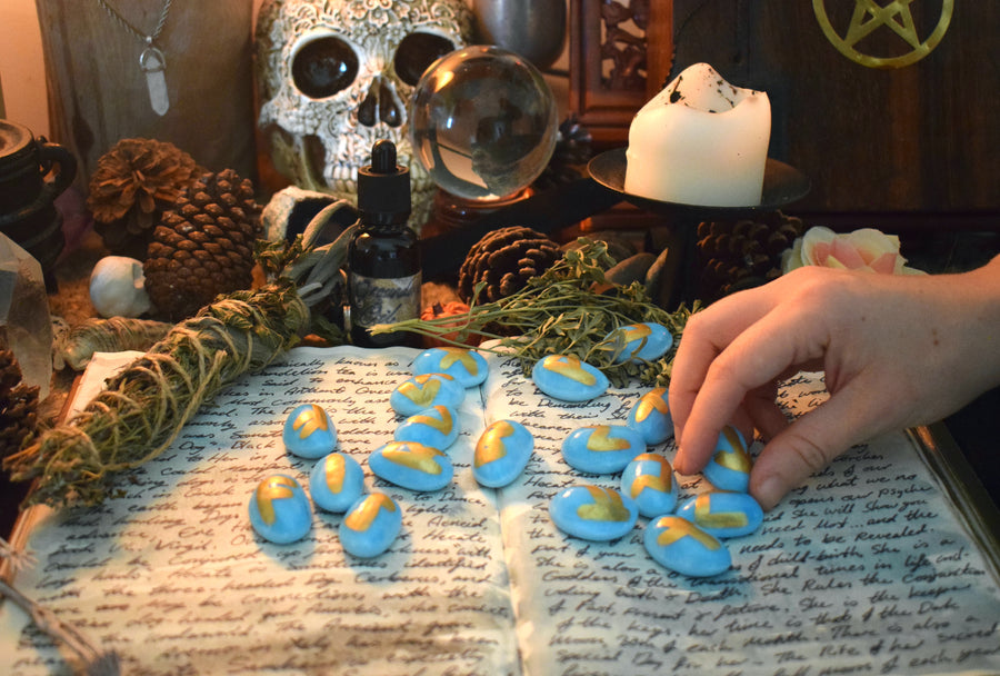 A set of handmade blue elder futhark runes resting on a grimoire surrounded by herbs, candles, crystals on an altar with a hand picking up one rune