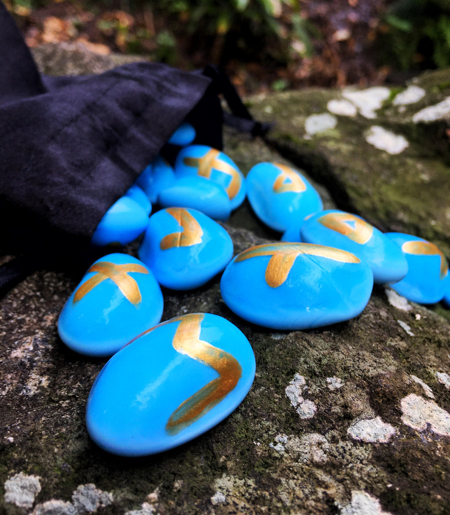 A set of handmade blue elder futhark runes spilling out of a black bag onto a mossy rock