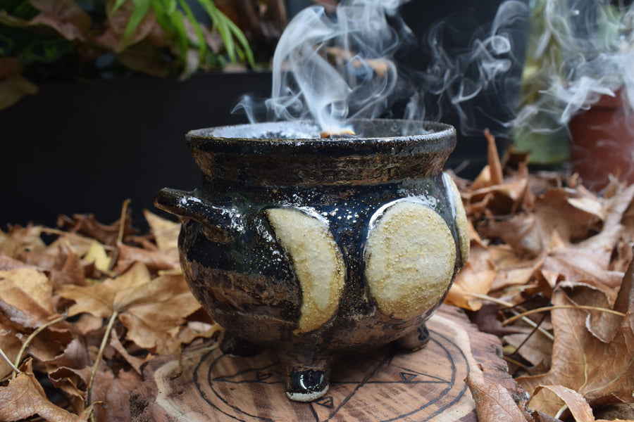 A cauldron designed with a triple moon symbol has smoke drifting from it while resting on a wood disk with a pentagram on it