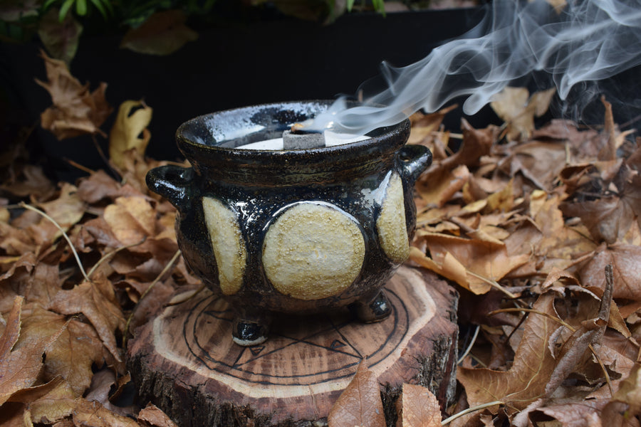 A cauldron designed with a triple moon symbol with smoke drifting from it resting on a wood disk with a pentagram on it on a bed of leaves