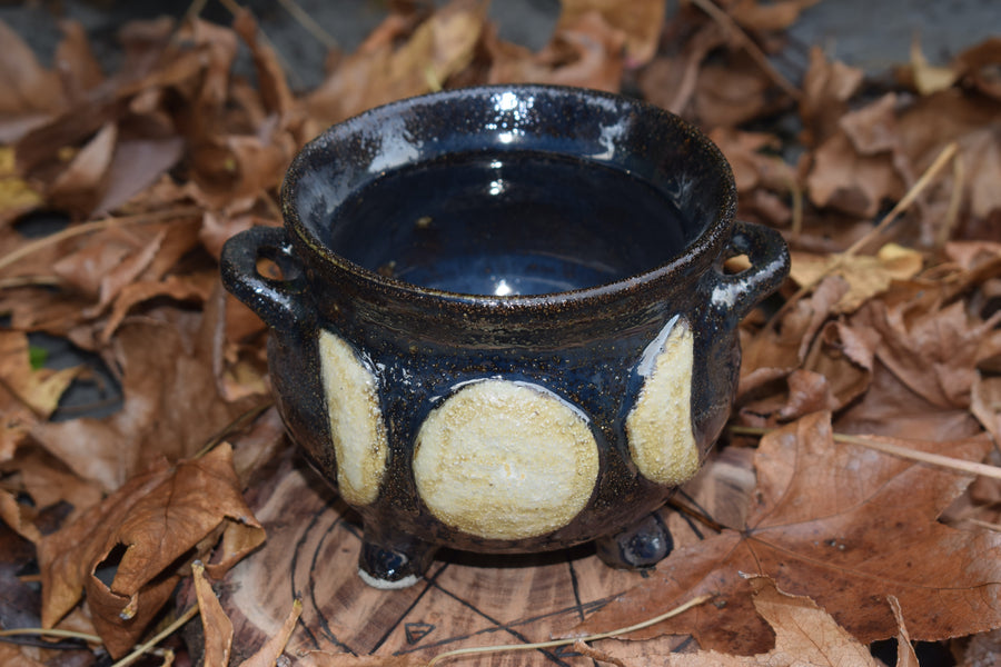 A cauldron designed with a triple moon symbol resting on a wood disk with a pentagram on it on a bed of leaves