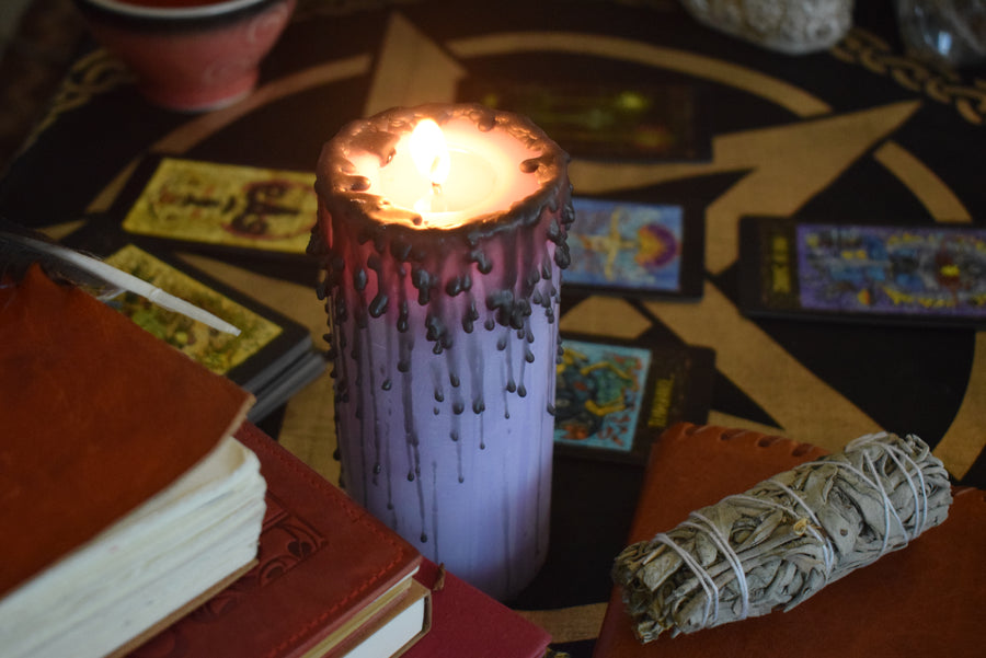 A purple pillar candle with black drips burns on an altar with a pentagram altar cloth, smudge stick, tarot cards and books