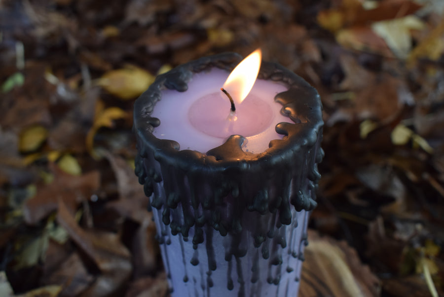 A purple pillar candle with black drips burns on a wooden disk with autumn leaves in the background