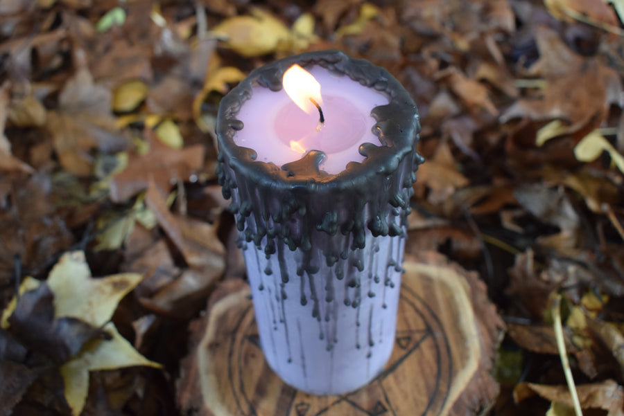 A purple pillar candle with black drips burns on a wooden disk with autumn leaves in the background