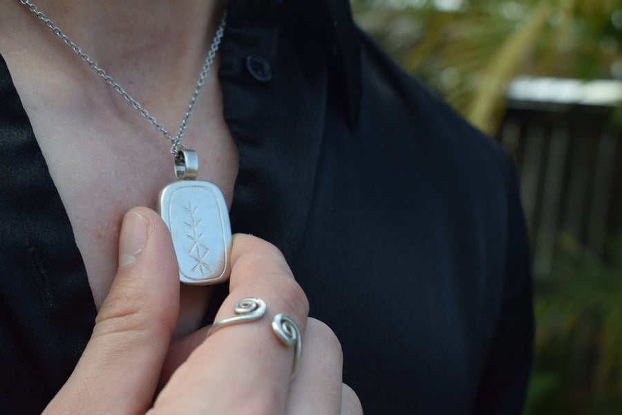 Person's neck with open shirt and hand showing handmade sterling silver pendant on chain inscribed with rune of health