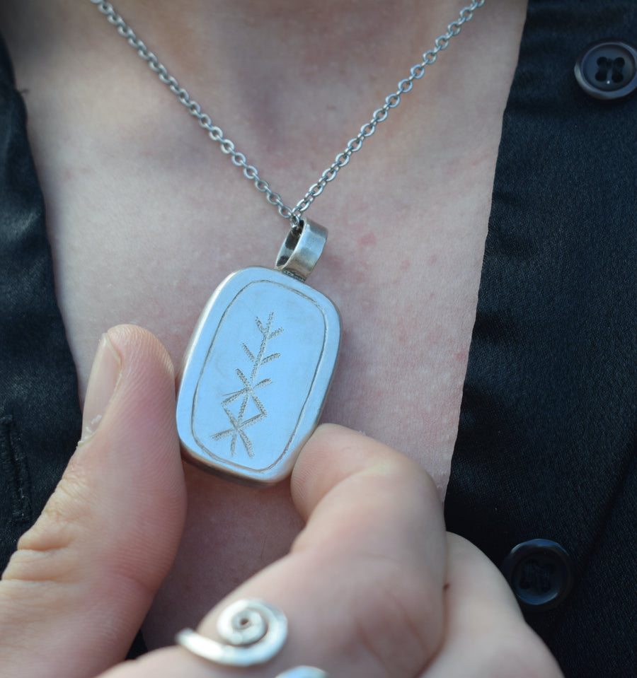 Person's neck with open shirt and hand showing handmade sterling silver pendant on chain inscribed with rune of health