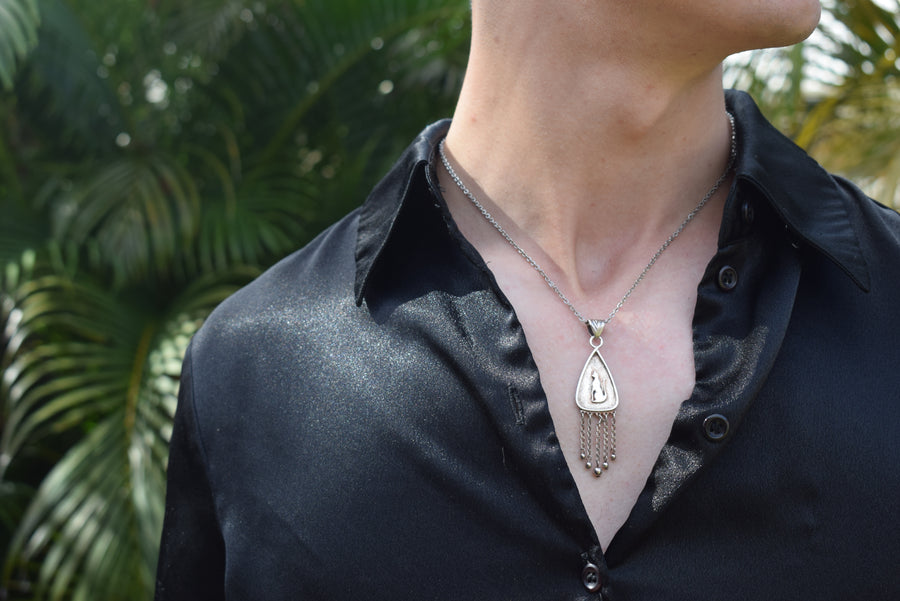 Neck of person wearing black shirt with sterling silver cat pendant with fringing necklace with greenery background