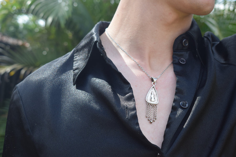 Neck of person wearing black shirt with sterling silver cat pendant with fringing necklace with background of greenery